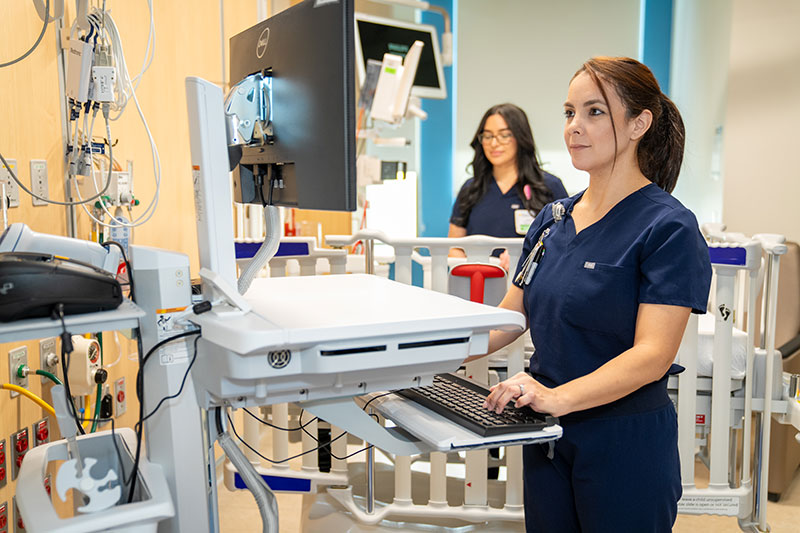 Two healthcare professionals in navy scrubs work at computer stations in a medical facility, with medical equipment visible in the background.